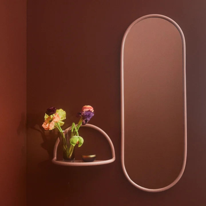 Der Angui Wandspiegel mit rosefarbenem Rahmen von AYTM neben einem kleinen Wandregal in derselben Farbe