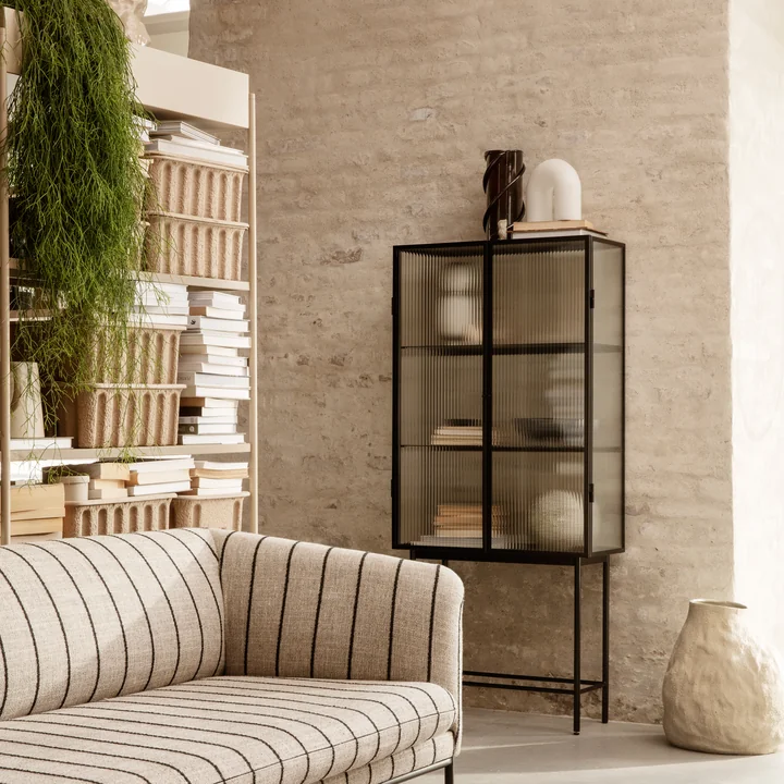 Die Haze Vitrine von Ferm Living mit vielen Kunstvasen und Büchern an einer Steinwand im Wohnzimmer