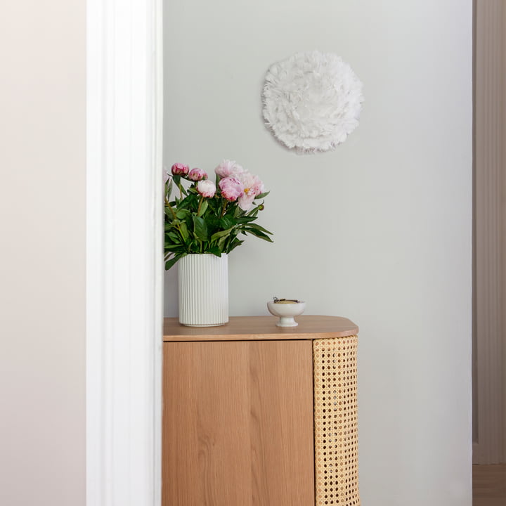 Die EOS Up Decken- und Wandleuchte Mini von Umage in créme über einem Holz-Sideboard und Blumen